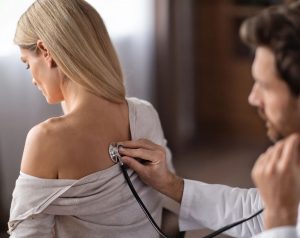 Médico neumólogo ausculta con un estetoscopio los pulmones de una paciente de mediana edad sentada en el consultorio, por posibles indicios de neumonía.