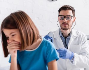 Niña pequeña con bata médica sentada en una camilla de consultorio mientras un médico la examina; la menor tose, posible signo de neumonía.