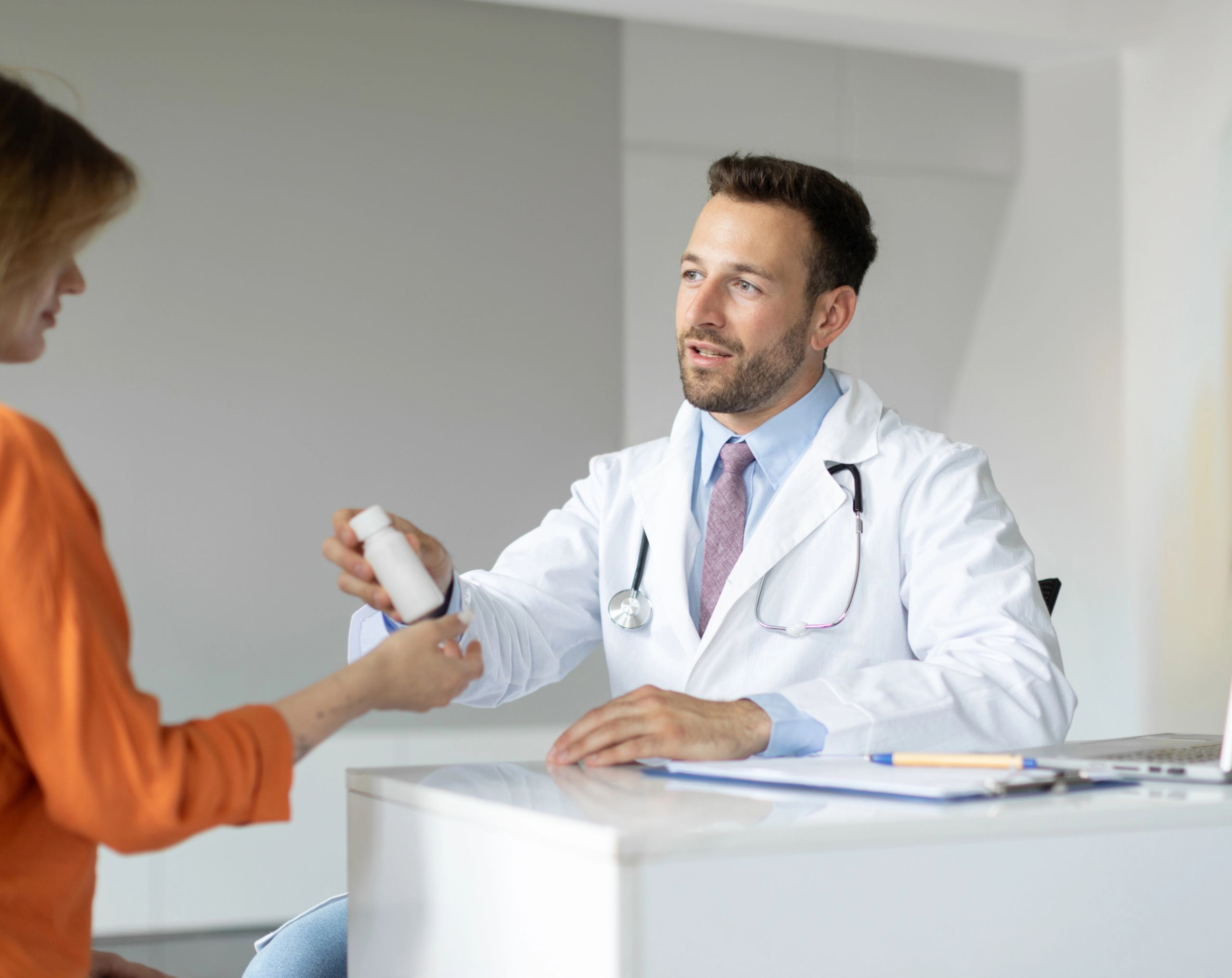 Médico joven con estetoscopio entrega un frasco de tratamiento antirretrovírico a una paciente mujer durante una consulta para el manejo oportuno del VIH.