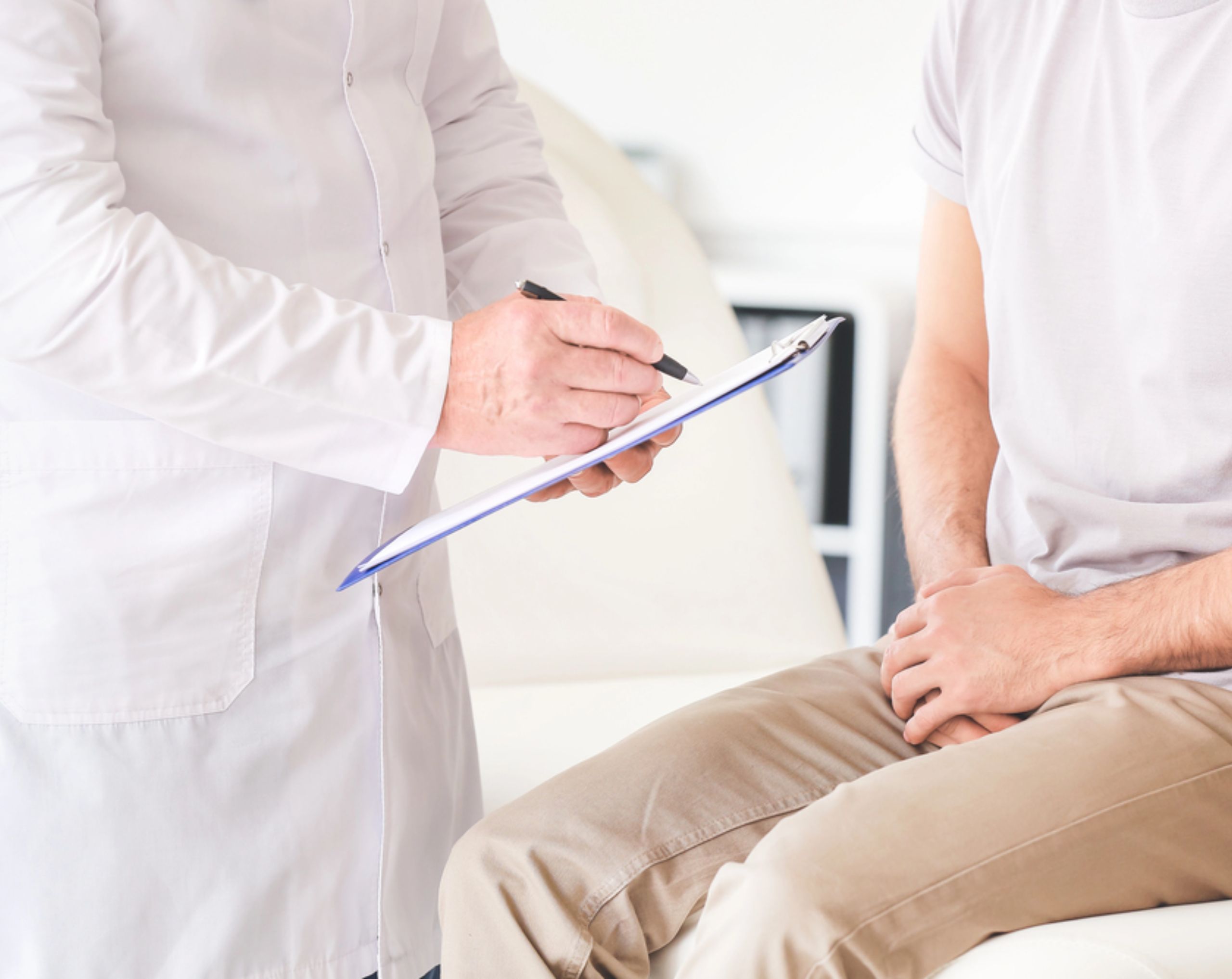 Hombre joven sentado en una camilla mientras un médico realiza una consulta, en representación del diagnóstico de la hiperplasia prostática benigna. 