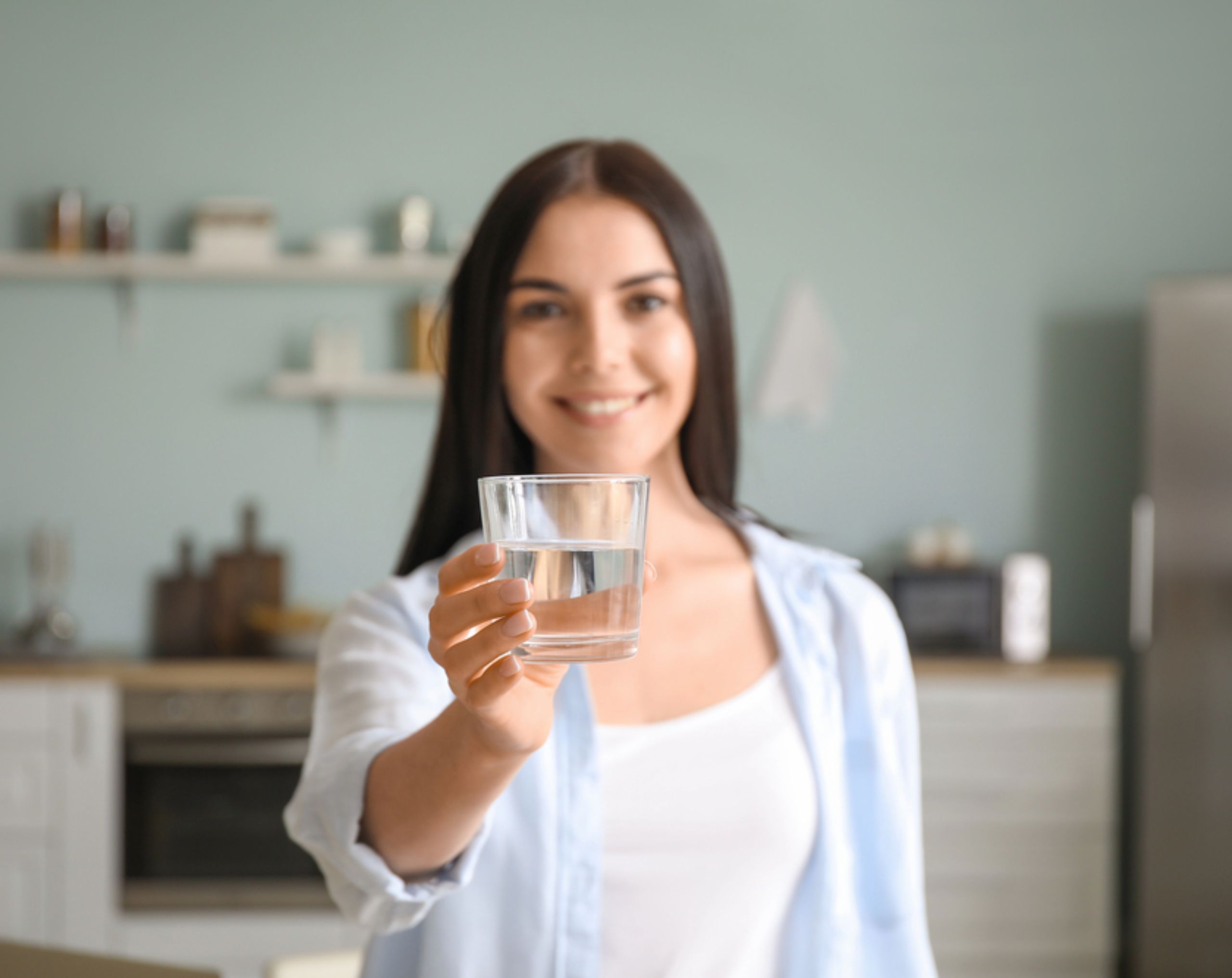 Mujer joven sonriente sostiene un vaso de agua hacia el frente, en representación de la hidratación como parte del manejo de la presión baja.