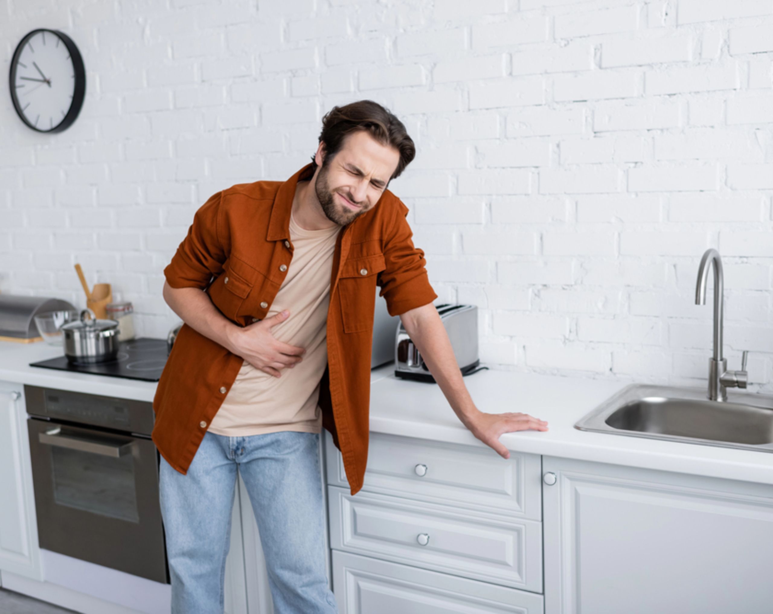 Hombre joven con camisa roja apoyado en la barra de la cocina mientras se sostiene el abdomen por dolor abdominal, uno de los síntomas de salmonella.