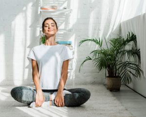 Mujer joven sentada en posición de flor de loto en su habitación practica respiración para controlar los síntomas del síndrome premenstrual.