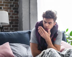 Hombre de mediana edad sentado en un sofá, tosiendo de forma persistente como síntoma de tuberculosis pulmonar.