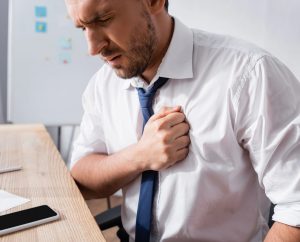 Hombre oficinista sentado frente a su escritorio se lleva la mano derecha al pecho debido a dolor, un síntoma de ataque al corazón.