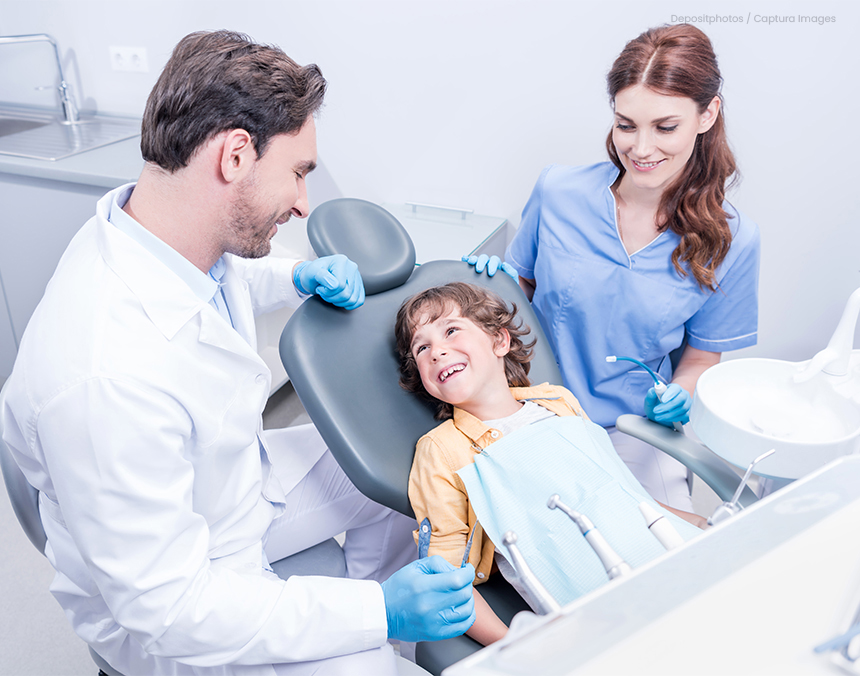 Niño sentado en una silla de dentista de color gris y sonriendo a un médico que usa bata blanca. Su madre se encuentra a un lado