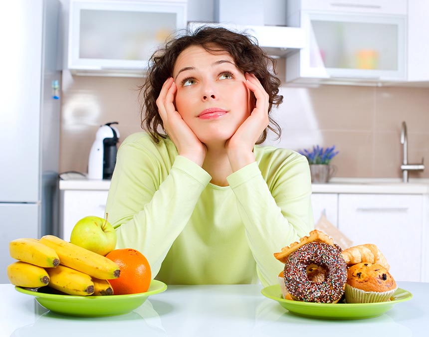 Mujer sentada en la silla de una cocina, con dos platos frente de ella, uno de fruta y otro de pan.