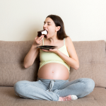 Mujer embarazada sentada en un sillón café, comiendo una rebanada de pastel.