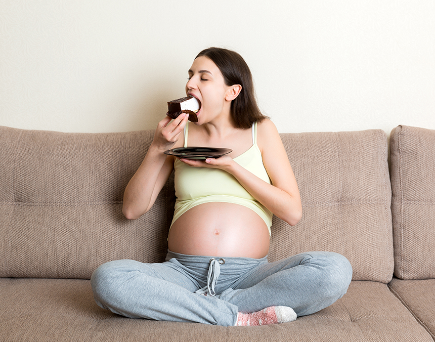Mujer embarazada sentada en un sillón café, comiendo una rebanada de pastel.