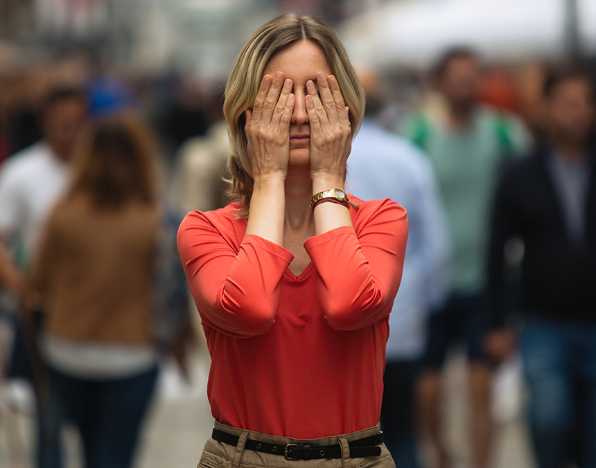 Mujer con playera roja y pantalón café, tapándose los ojos y el fondo difuminado.