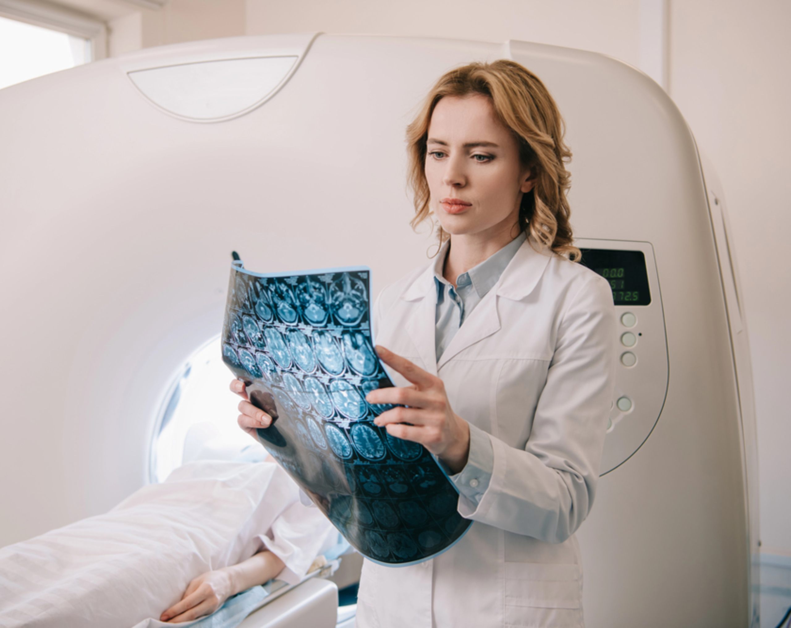 Médica sostiene una imagen de resonancia magnética cerebral mientras la paciente permanece recostada en el resonador.