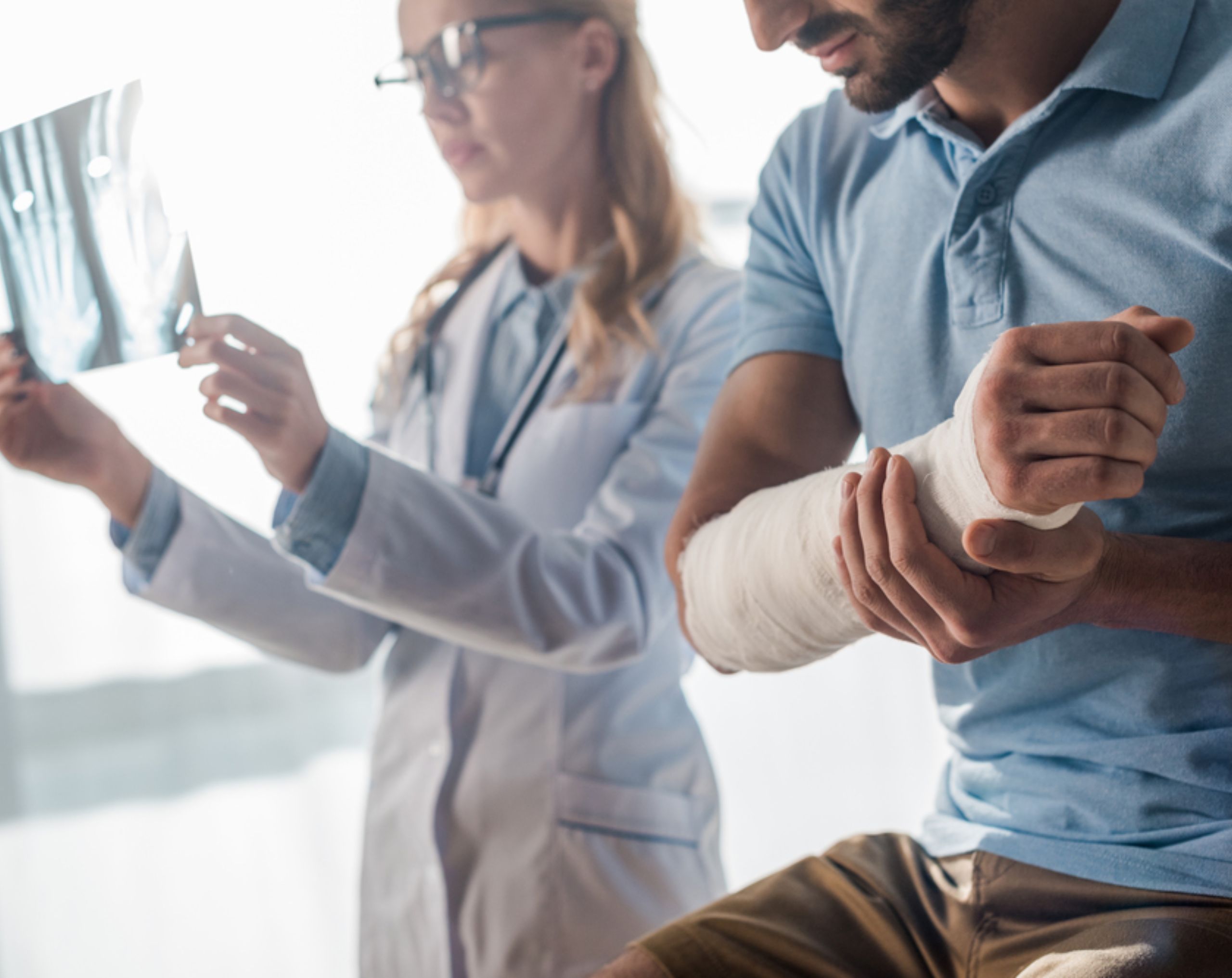 Hombre de mediana edad con fractura en el brazo sentado en consulta mientras una doctora revisa su radiografía para evaluar el tratamiento.