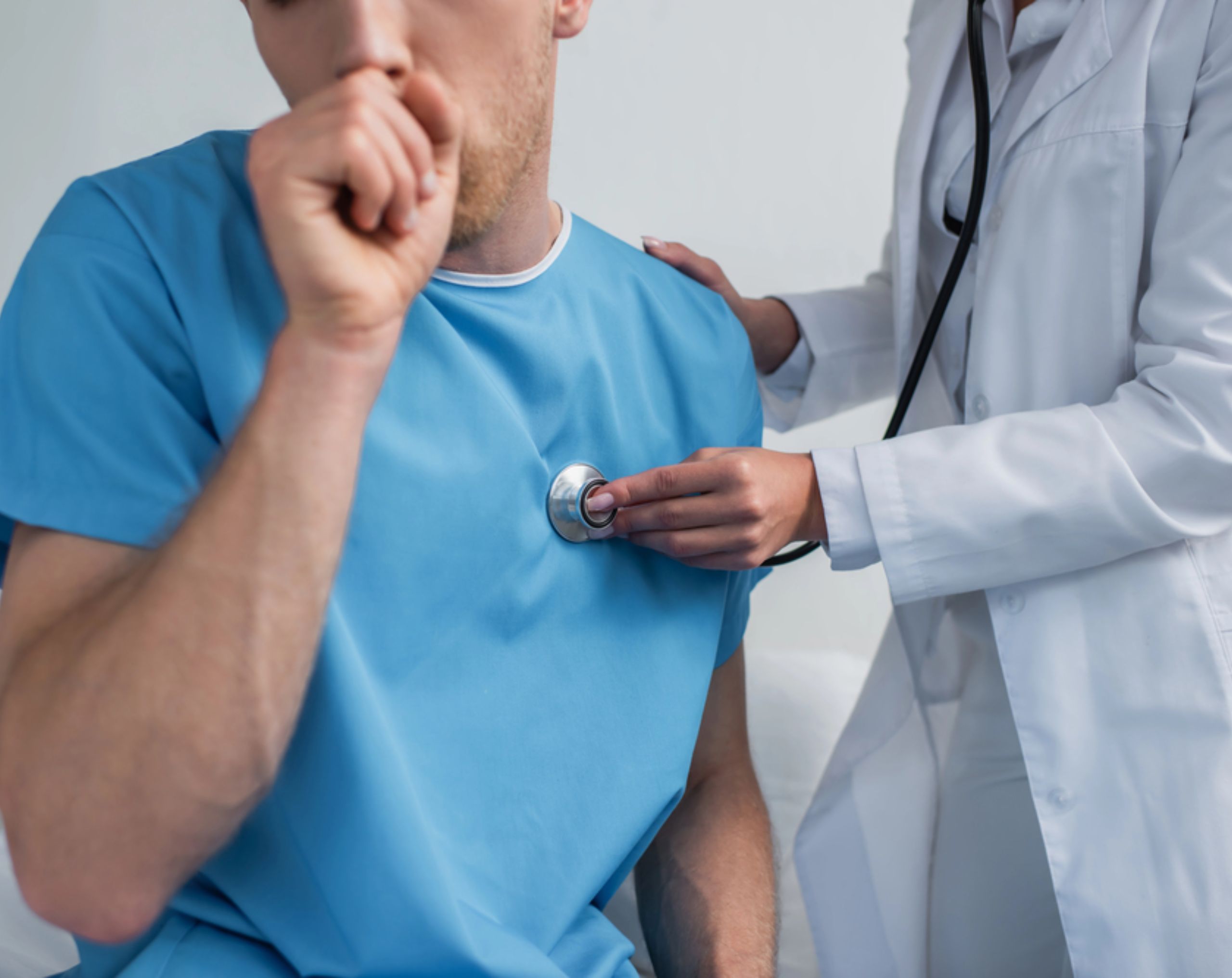Paciente masculino sentado en una camilla, tosiendo y cubriéndose la boca, mientras una doctora escucha sus pulmones con un estetoscopio ante posible neumonía, una enfermedad nosocomial.