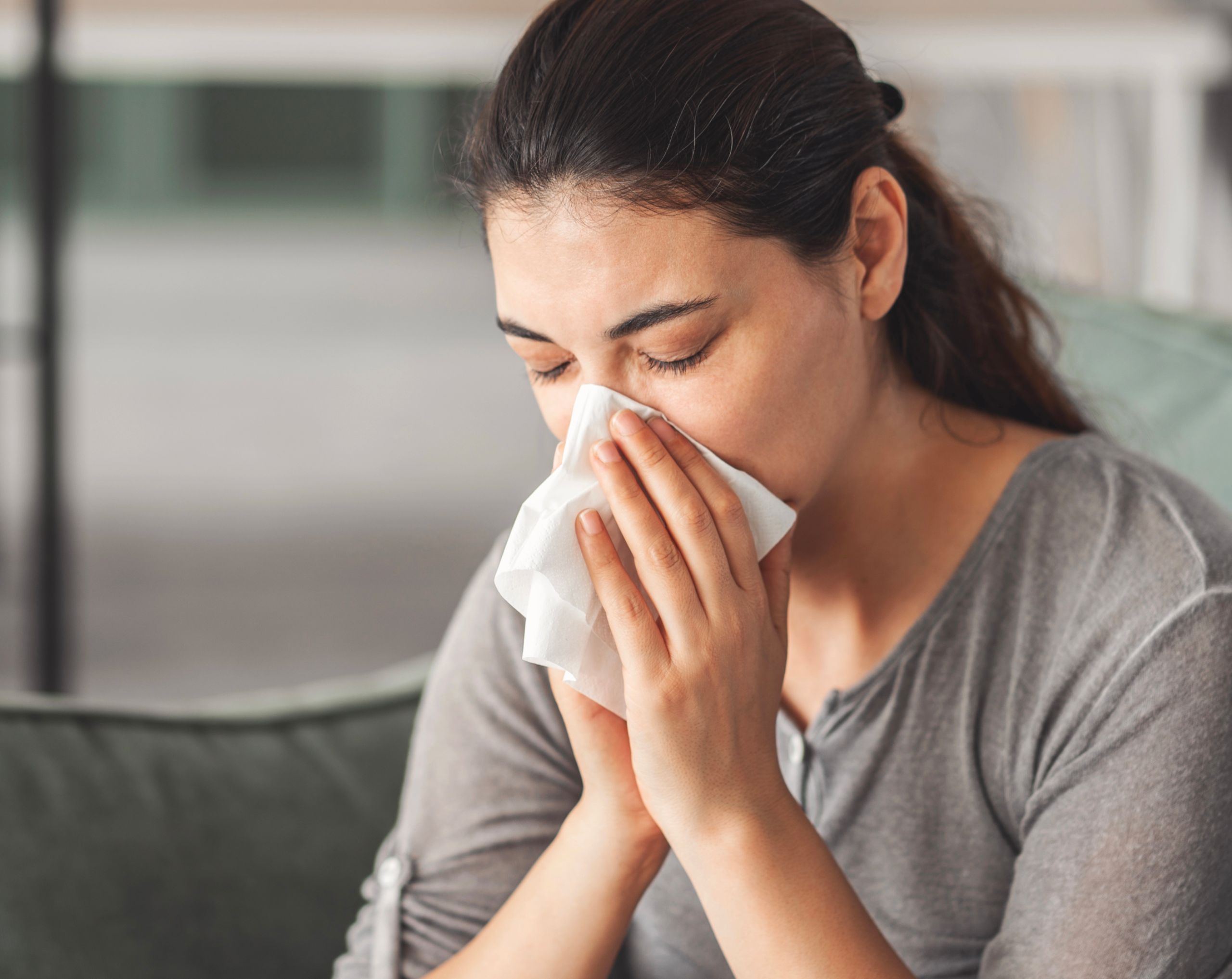 Mujer joven sentada en un sillón, limpiándose la nariz por goteo postnasal, un síntoma común de sinusitis.