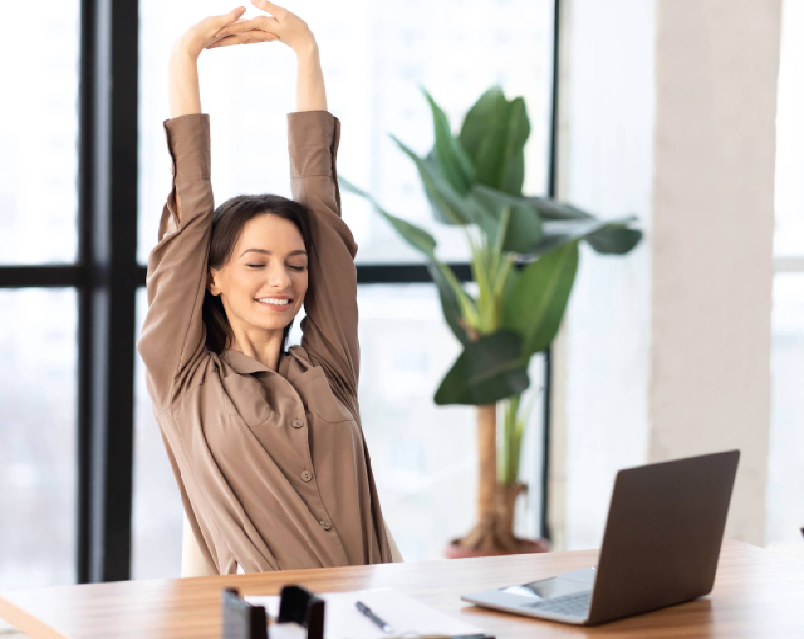 Mujer joven hace pausa activa en la oficina estirando los brazos por arriba de la cabeza, frente a un escritorio con una computadora.
