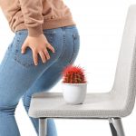 Mujer a punto de sentarse en una silla con un cactus, sosteniendo la zona del ano con una mano, en representación del ardor y comezón causados por las almorranas.