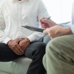 Paciente masculino de mediana edad en consultorio médico, mostrando molestias en la zona testicular mientras el urólogo explica diagnóstico por posible cáncer de testículo.