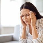 Joven mujer sentada en un sofá, sosteniendo con ambas manos las sienes mientras sufre mareos y fatiga, síntomas de disautonomía.