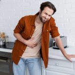 Hombre de mediana edad en la cocina, apoyado en el mueble mientras sostiene el abdomen con gesto de dolor por síntomas de diverticulitis.