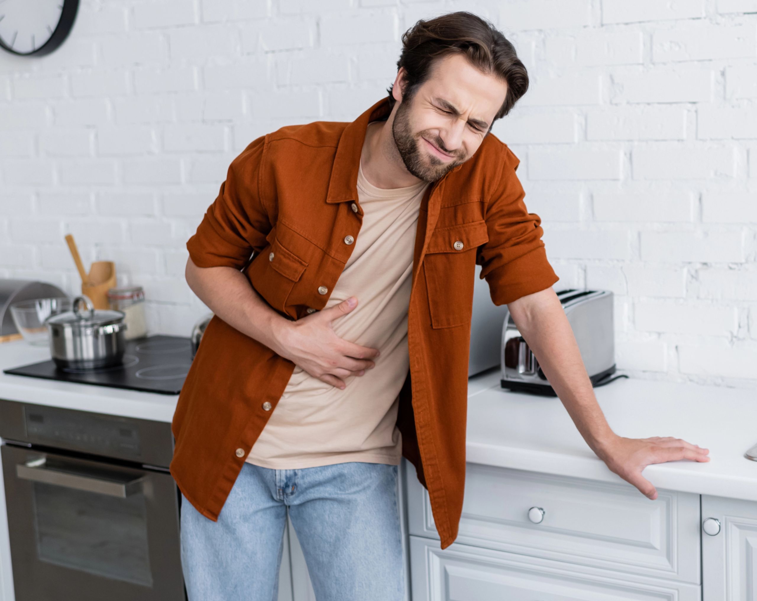 Hombre de mediana edad en la cocina, apoyado en el mueble mientras sostiene el abdomen con gesto de dolor por síntomas de diverticulitis.
