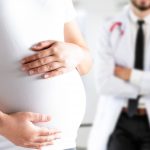 Mujer embarazada sostiene su abdomen con ambas manos como señal de posible embarazo de alto riesgo; al fondo, un médico observa.