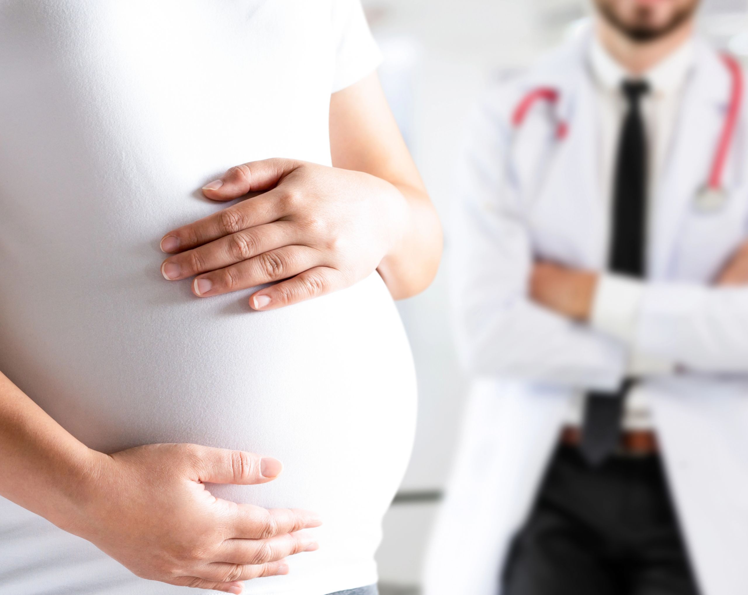 Mujer embarazada sostiene su abdomen con ambas manos como señal de posible embarazo de alto riesgo; al fondo, un médico observa.