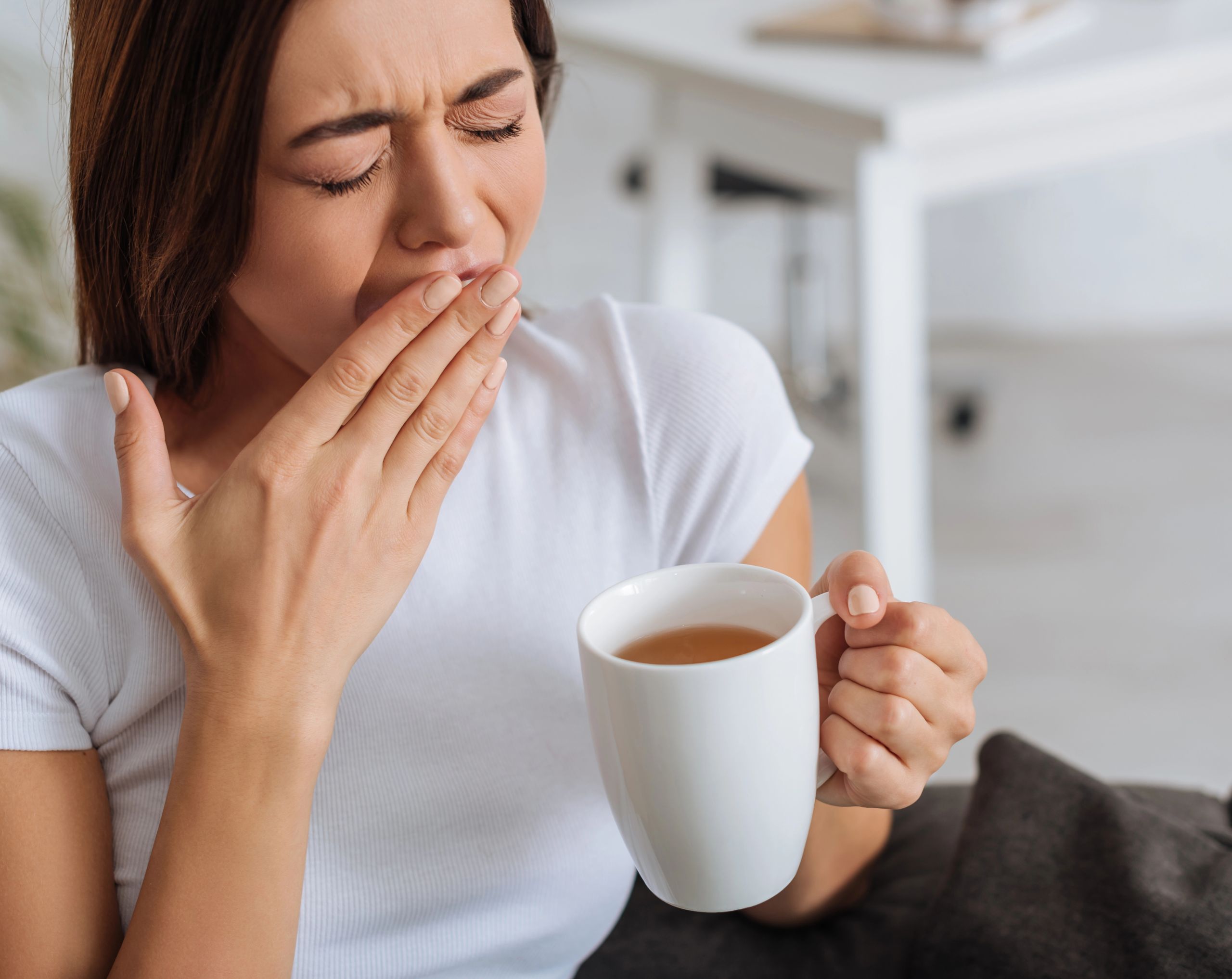 Mujer de mediana edad sosteniendo una taza de café y bostezando, mostrando somnolencia, un posible síntoma de Tiroiditis de Hashimoto.
