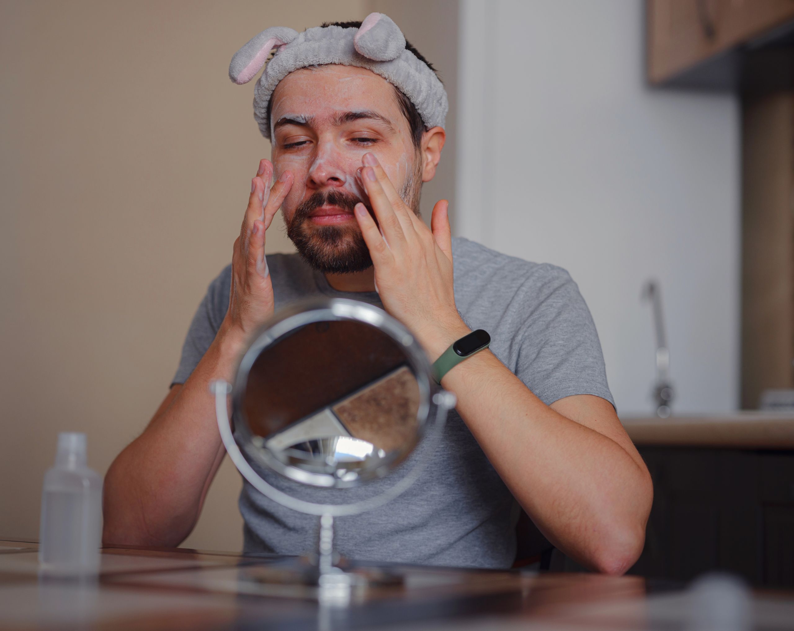 Hombre joven sentado frente al espejo aplicando una crema en su rostro como tratamiento para el acné hormonal.