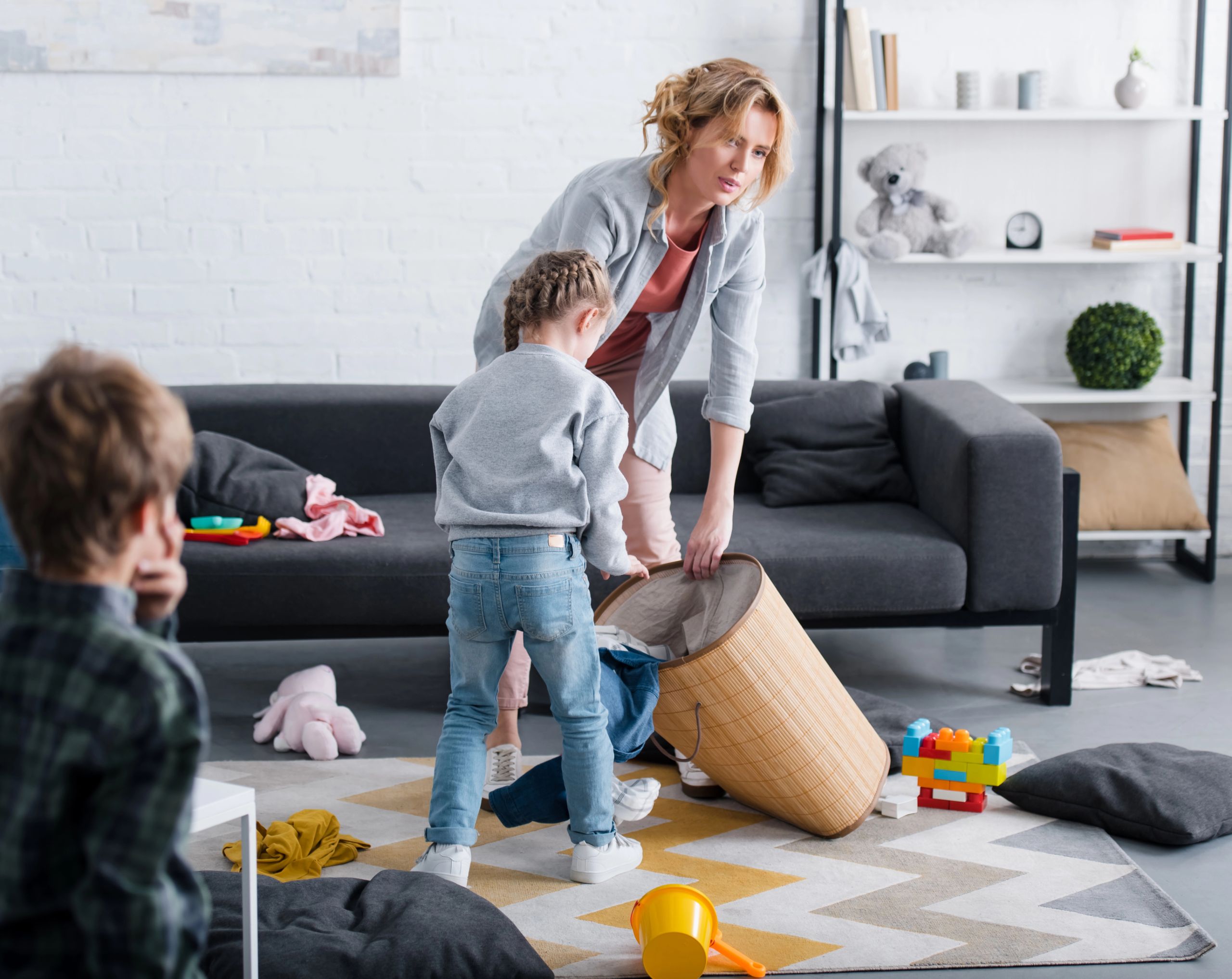 Mujer adulta intentando recoger los juguetes esparcidos por el piso con una cesta, mientras sus hijos corren alrededor; la escena refleja hiperactividad, posible síntoma de TDAH