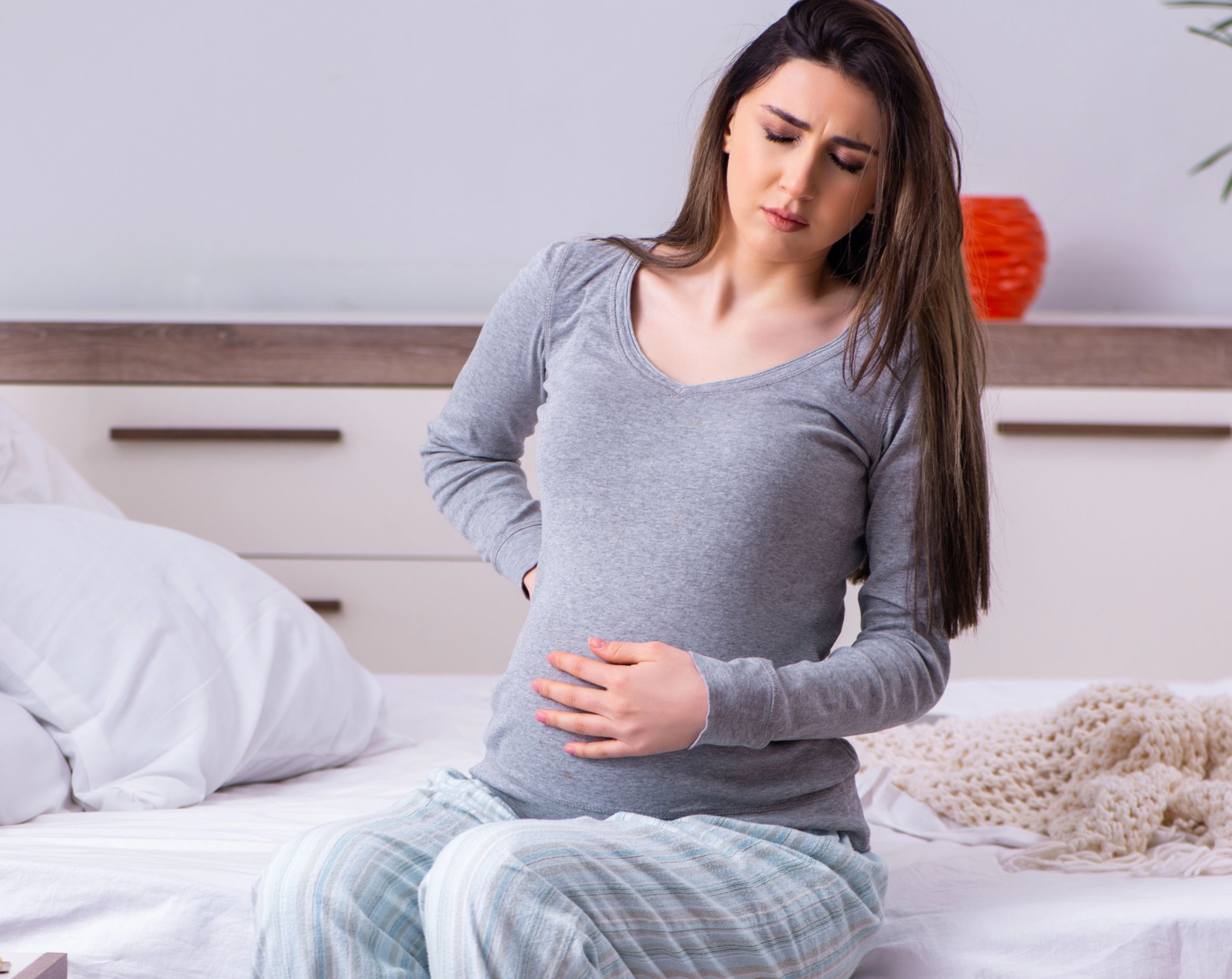 Mujer embarazada sentada en su cama sosteniendo su pancita con una mano mostrando molestias por contracciones de Braxton Hicks. 