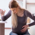 Mujer joven con 30 semanas de embarazo sentada en el comedor, muestra una leve expresión de dolor por las contracciones de Braxton Hicks.