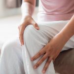 Mujer de mediana edad sentada en el sofá mientras sostiene con ambas manos la rodilla por dolor causado por el quiste de Baker.