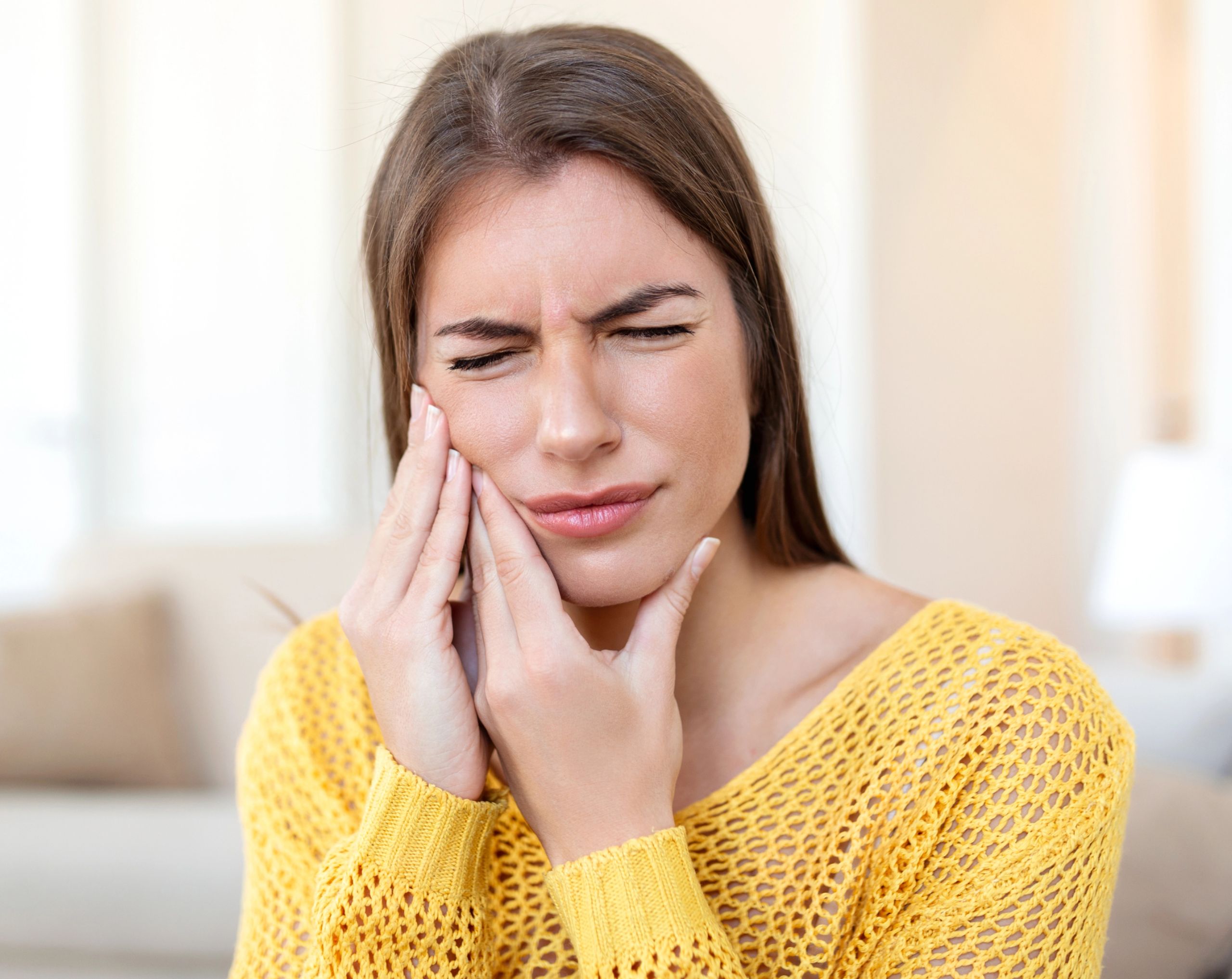 Mujer joven sostiene su mejilla y mandíbula con ambas manos debido a un dolor intenso causado por un absceso dental.
