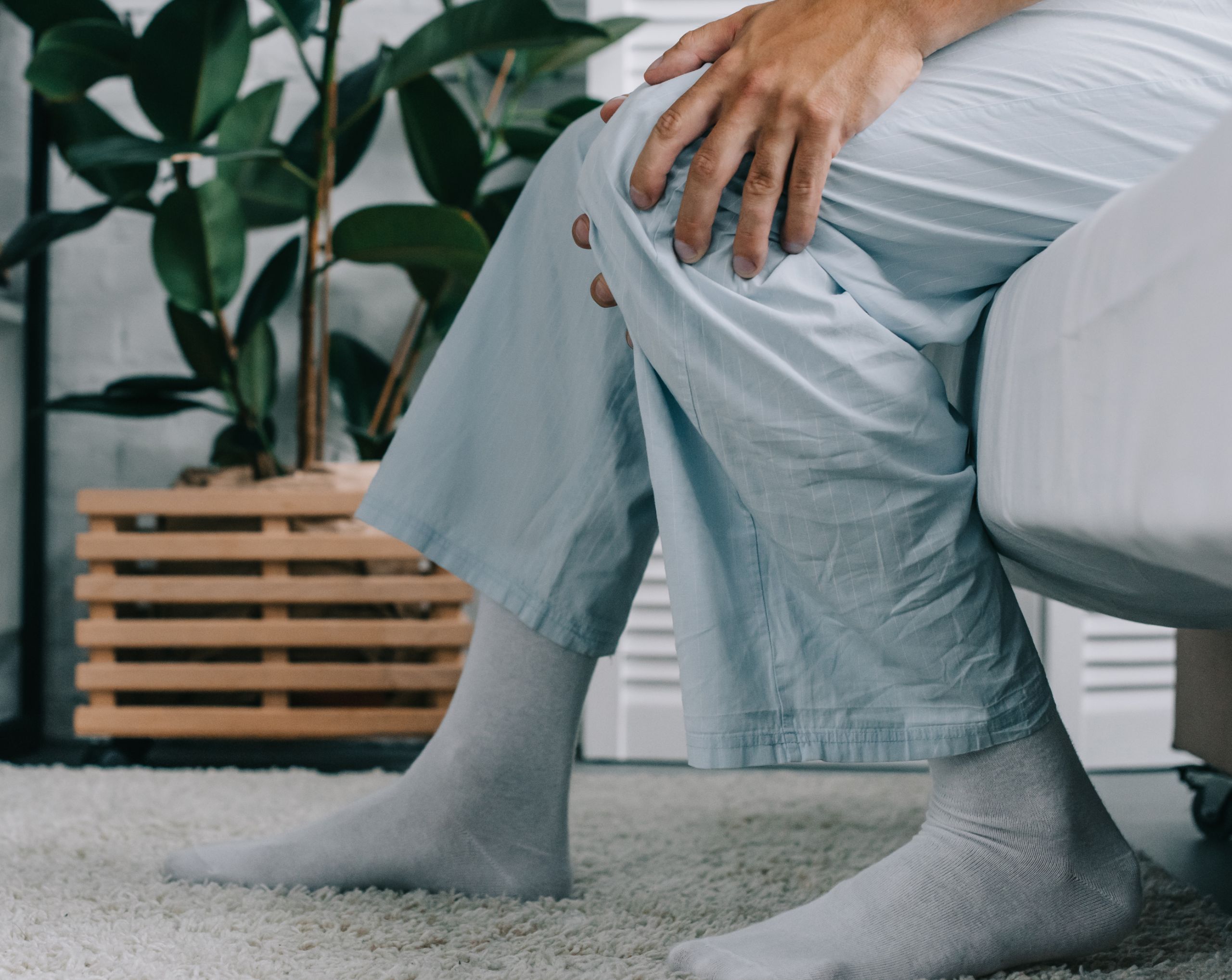 Hombre de mediana edad sentado en la cama con los pies apoyados en el suelo, sostiene con ambas manos la parte posterior de la rodilla debido al dolor causado por un quiste de Baker.
