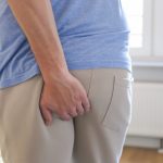 Hombre adulto mayor coloca su mano en la zona baja de la espalda debido al dolor provocado por un absceso perianal.