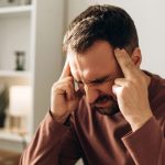 Hombre de mediana edad sentado en un sofá se sostiene las sienes con expresión de dolor intenso, signo de una posible hemorragia subaracnoidea.