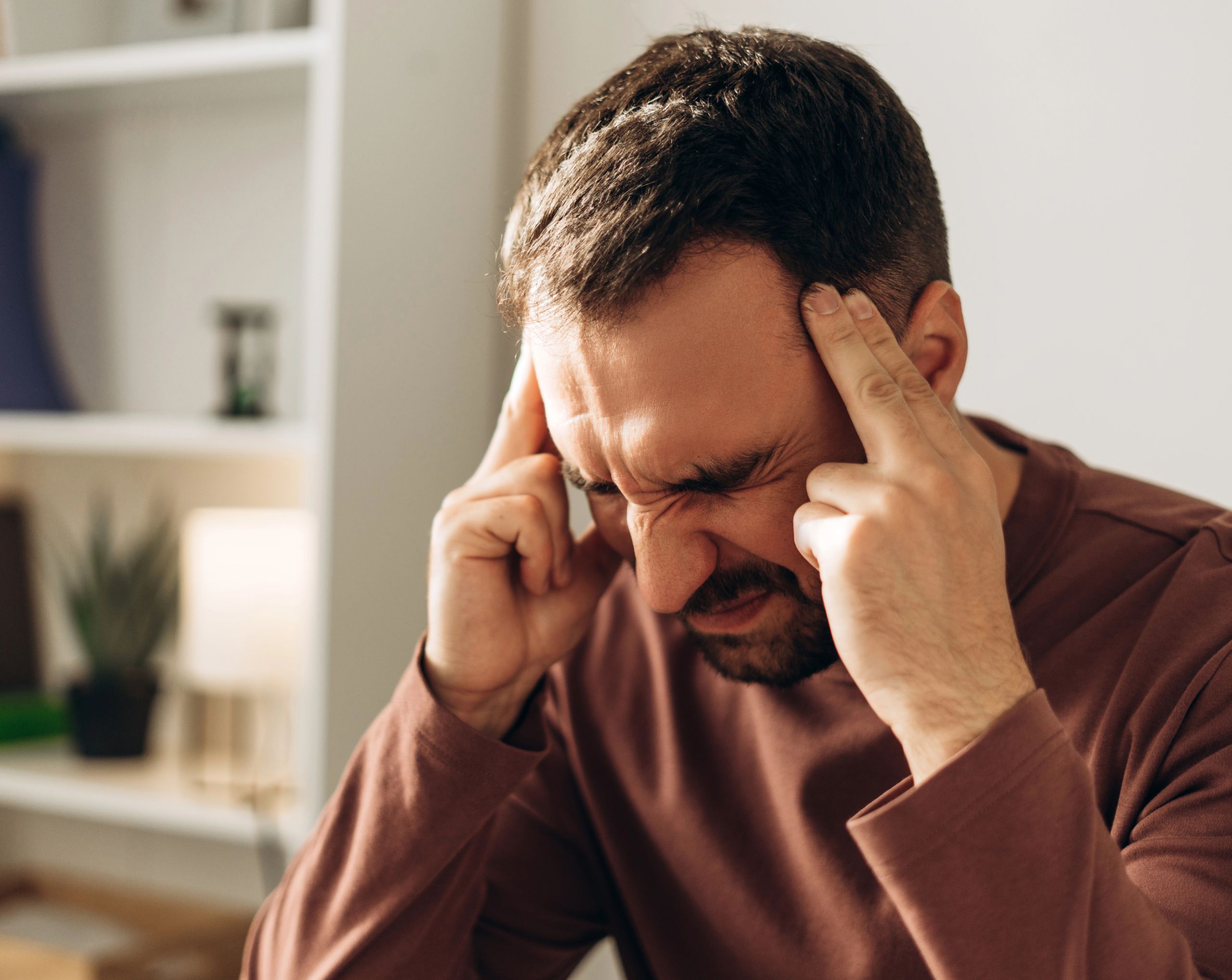 Hombre de mediana edad sentado en un sofá se sostiene las sienes con expresión de dolor intenso, signo de una posible hemorragia subaracnoidea.