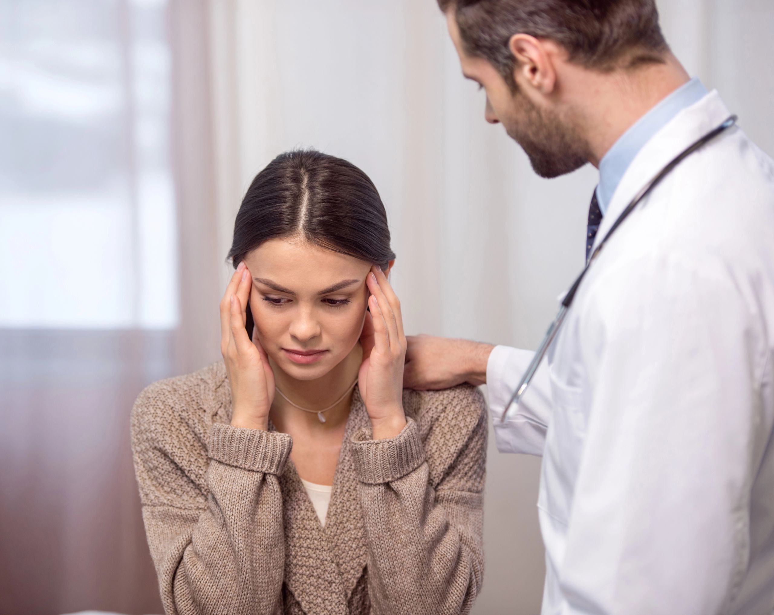 Mujer sentada en un consultorio se toca las sienes con ambas manos mientras un médico la revisa por un fuerte dolor de cabeza relacionado con una hemorragia subaracnoidea.