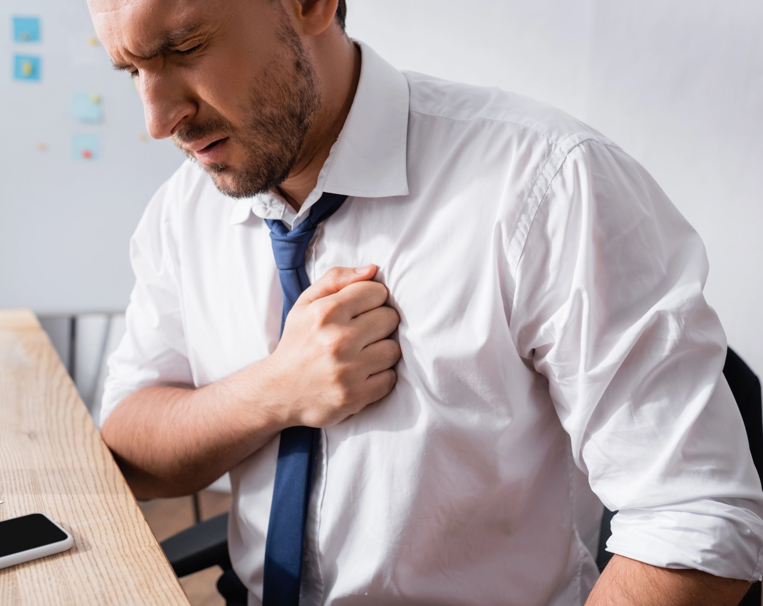 Hombre adulto en oficina, vestido con camisa formal y corbata, se toca el pecho con la mano derecha debido a doloren el pecho asociado a bradicardia sinusal.