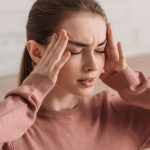 Mujer joven con dolor de cabeza intenso, presionando sus sienes, posible síntoma de meningitis.