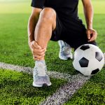 Hombre en cuclillas en cancha de fútbol sostiene su tobillo derecho por lesión durante el juego.