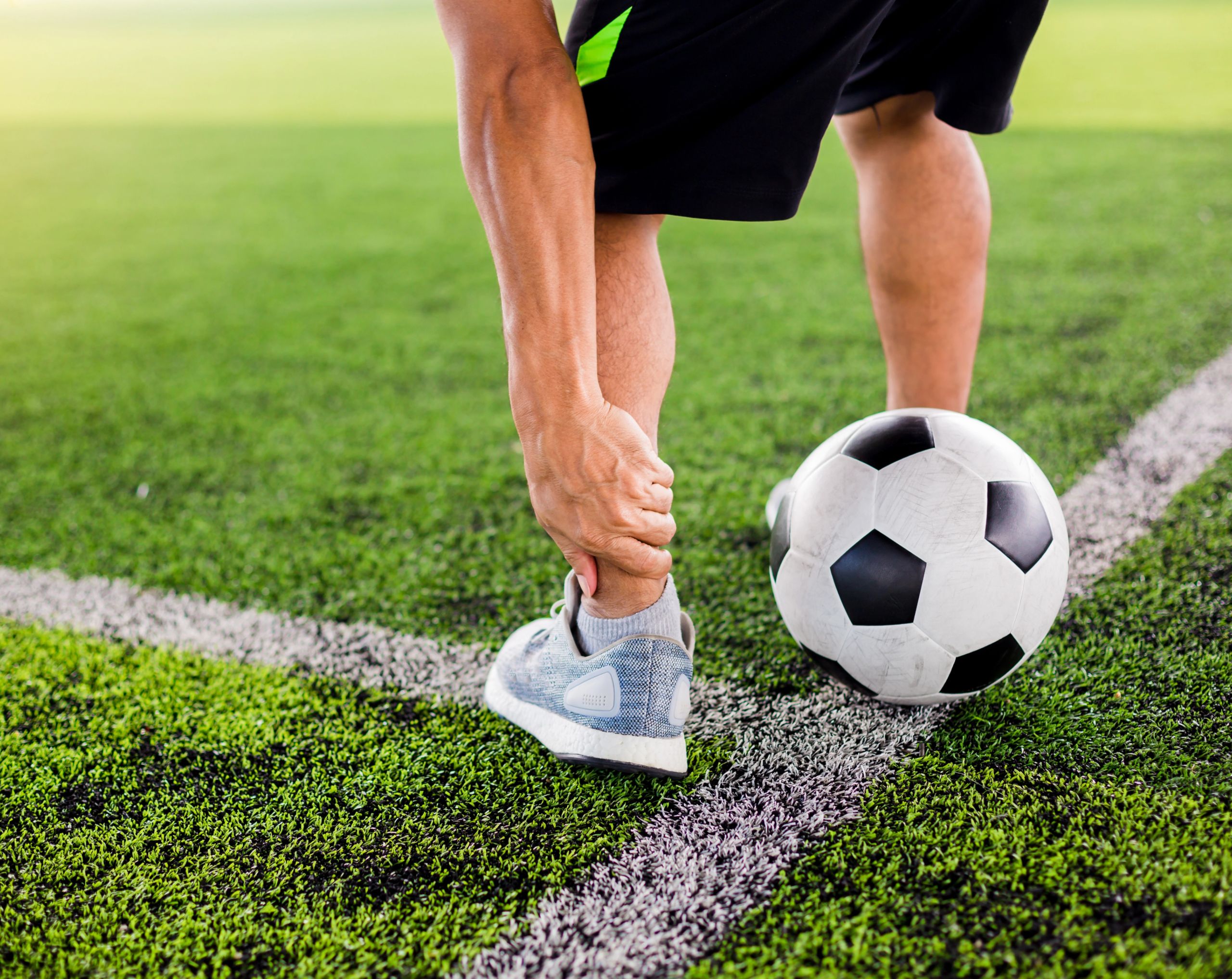 Hombre en cancha de fútbol sostiene su tobillo por posible lesión durante el juego.