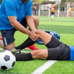 Entrenador deportivo revisando la lesión de rodilla de un futbolista recostado en la cancha durante un partido.