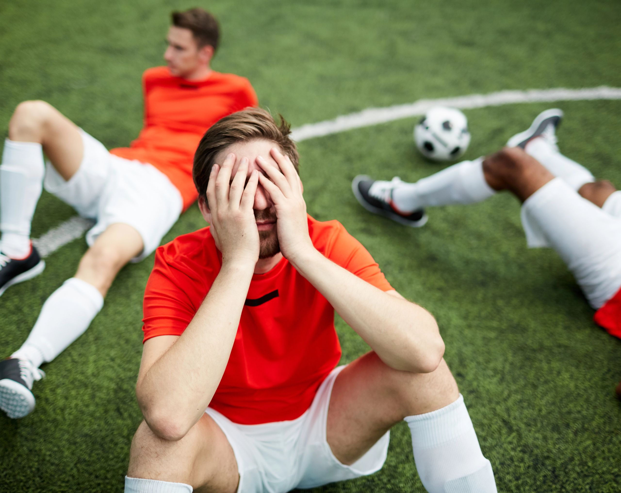 Hombre joven futbolista sentado en la cancha cubre su rostro con ambas manos en representación de la fatiga que siente, síntoma de muerte súbita cardíaca. 