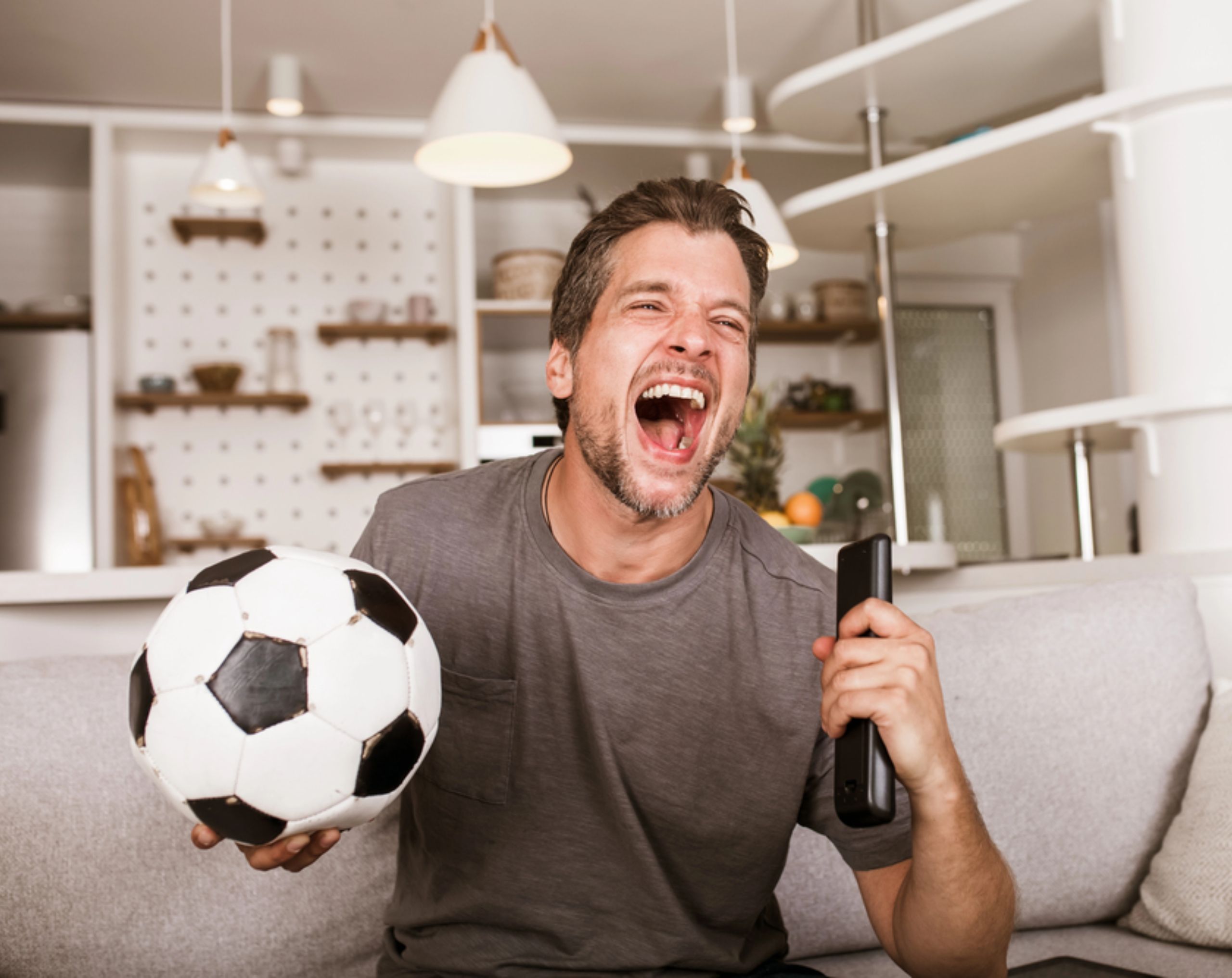 Hombre aficionado al futbol sentado en un sofá sosteniendo un balón mientras celebra con emoción durante un partido.
