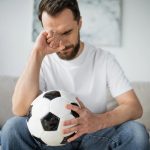 Hombre aficionado del futbol con expresión de tristeza sentado en la sala de su casa viendo un partido de futbol.