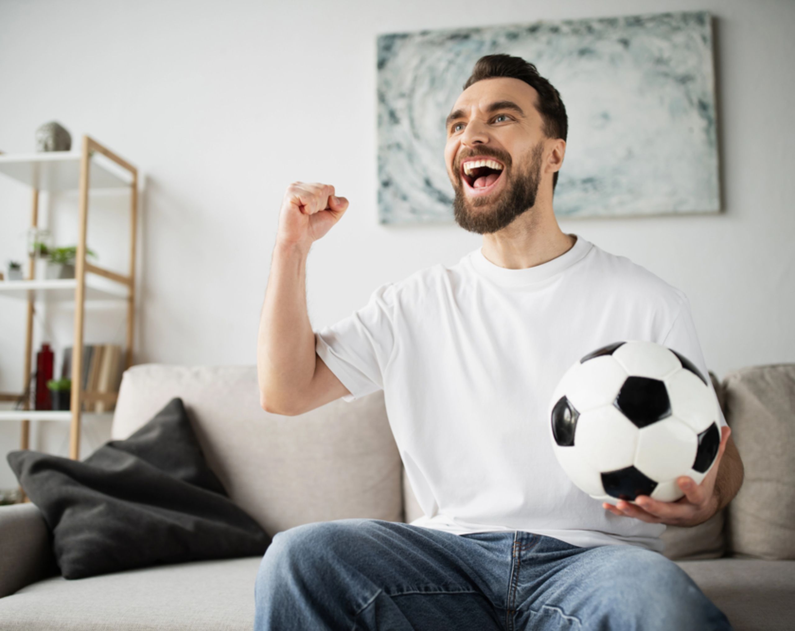 Aficionado del futbol celebra con emoción frente al televisor mientras ve un partido y sostiene un balón.