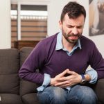 Hombre de mediana edad sentado en un sofá, sujetándose el abdomen con ambas manos debido a un dolor abdominal, uno de los síntomas de la ciclosporosis.