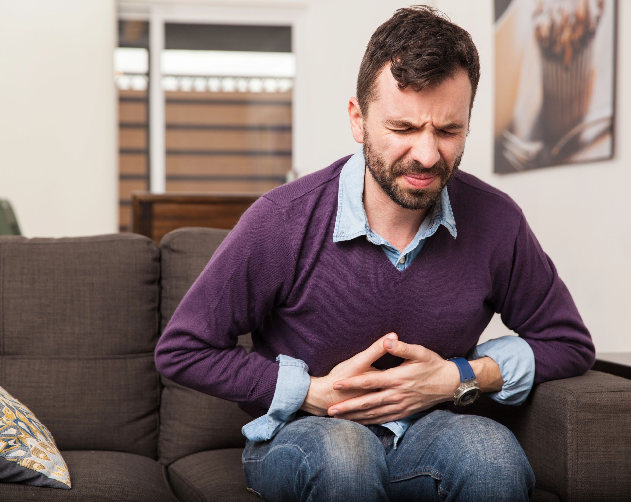 Hombre de mediana edad sentado en un sofá, sujetándose el abdomen con ambas manos debido a un dolor abdominal, uno de los síntomas de la ciclosporosis.
