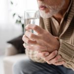 Hombre adulto mayor con Parkinson sostiene un vaso de agua con una mano mientras la otra la sujeta debido a los temblores.